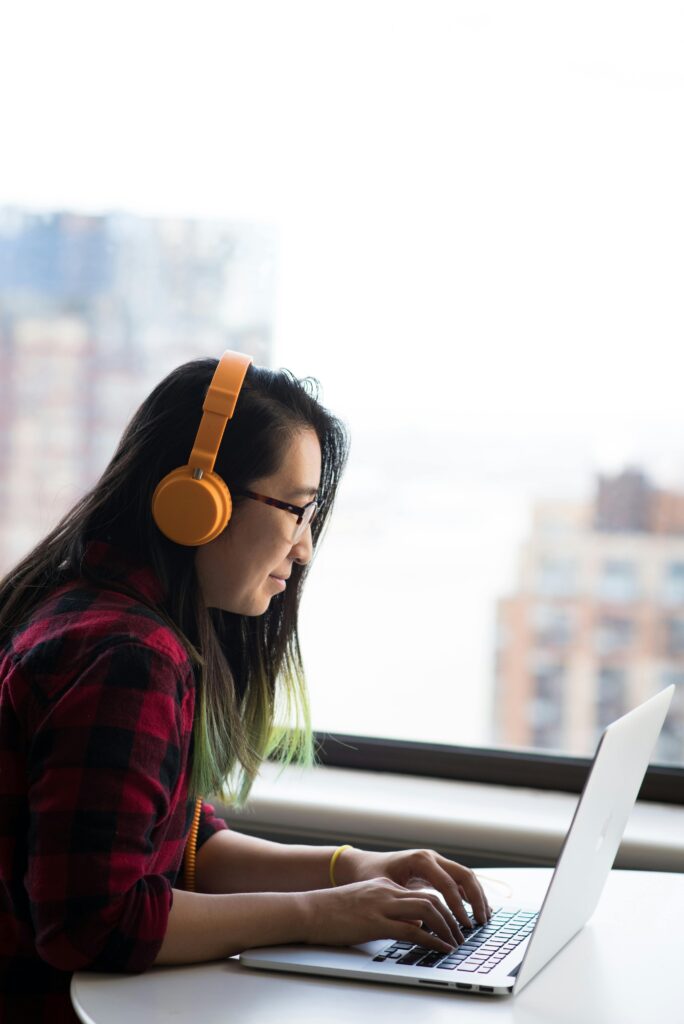 Asian woman in headphones working remotely on a laptop by the window, illustrating modern digital nomad lifestyle.